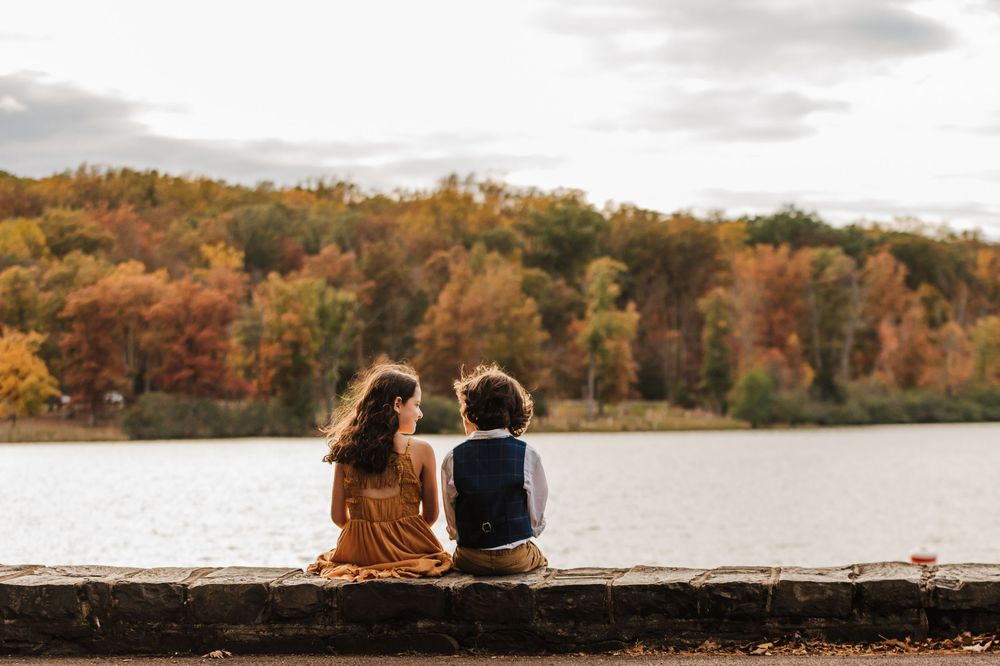 Couple sits together on lakeside dock surrounded by vibrant autumn foliage reflecting in calm waters.