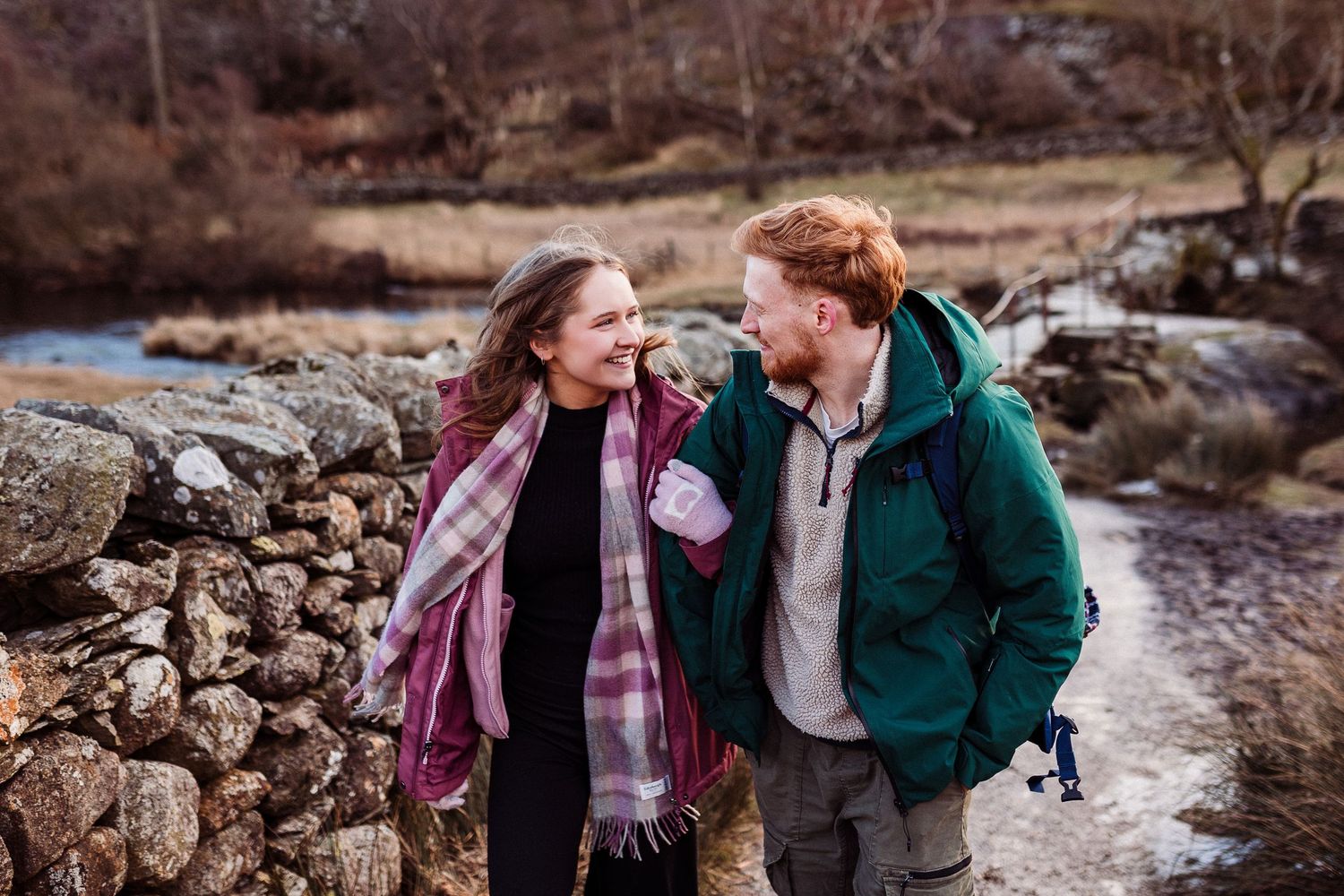 Two people laugh together while walking on a gravel path through scenic Scottish Highlands with snow-capped mountains.