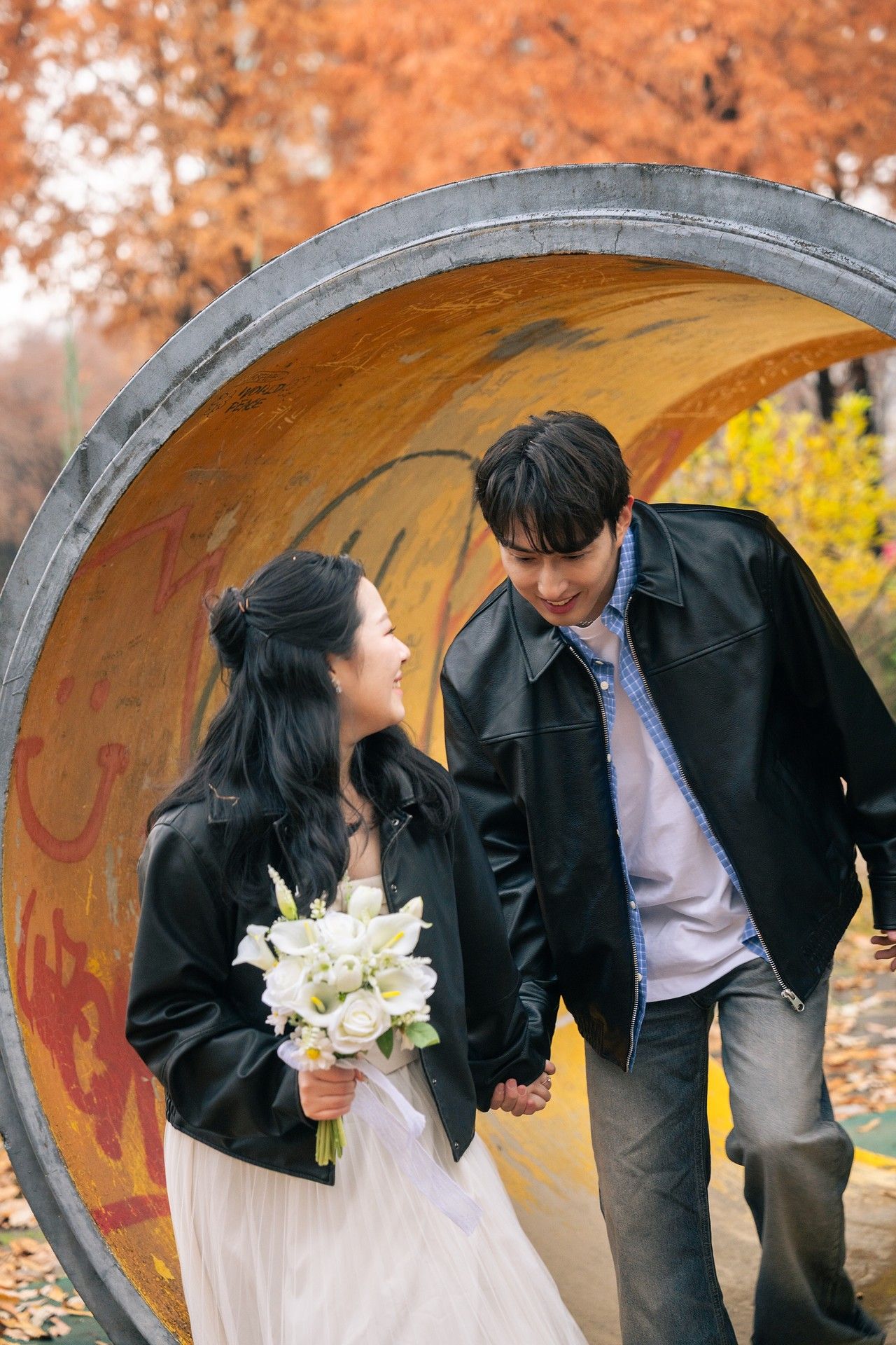 A couple shares a romantic moment in front of a circular tunnel during autumn, with the bride holding white flowers.