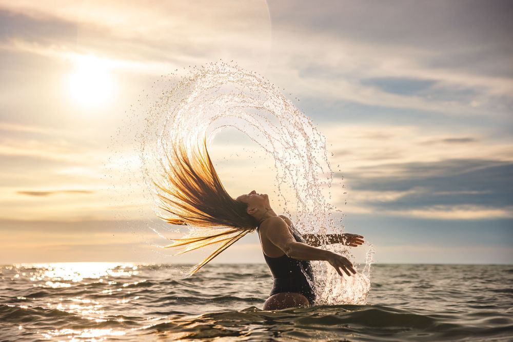 A dramatic hair flip creates a water arc against a golden sunset over the ocean.