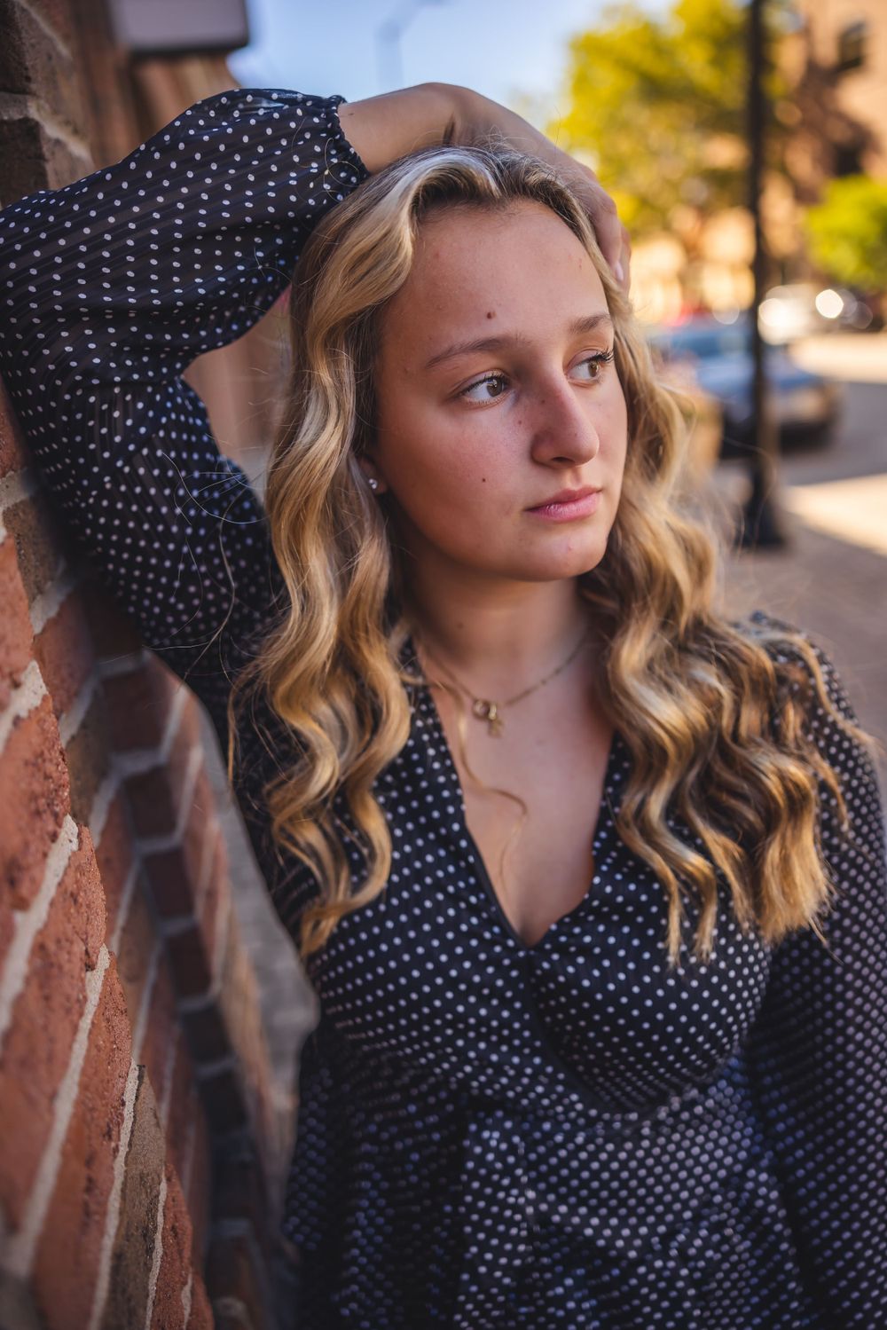 A young woman in polka dot dress poses against brick wall in warm afternoon light.