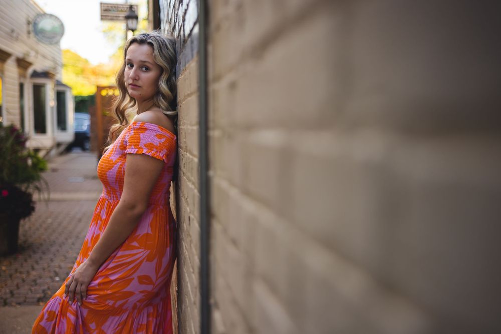 Woman in vibrant orange floral dress poses against textured wall in urban alleyway at golden hour.