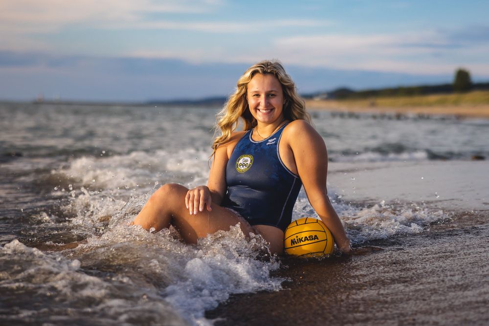 Water polo player sits in breaking waves with yellow ball at sunset.