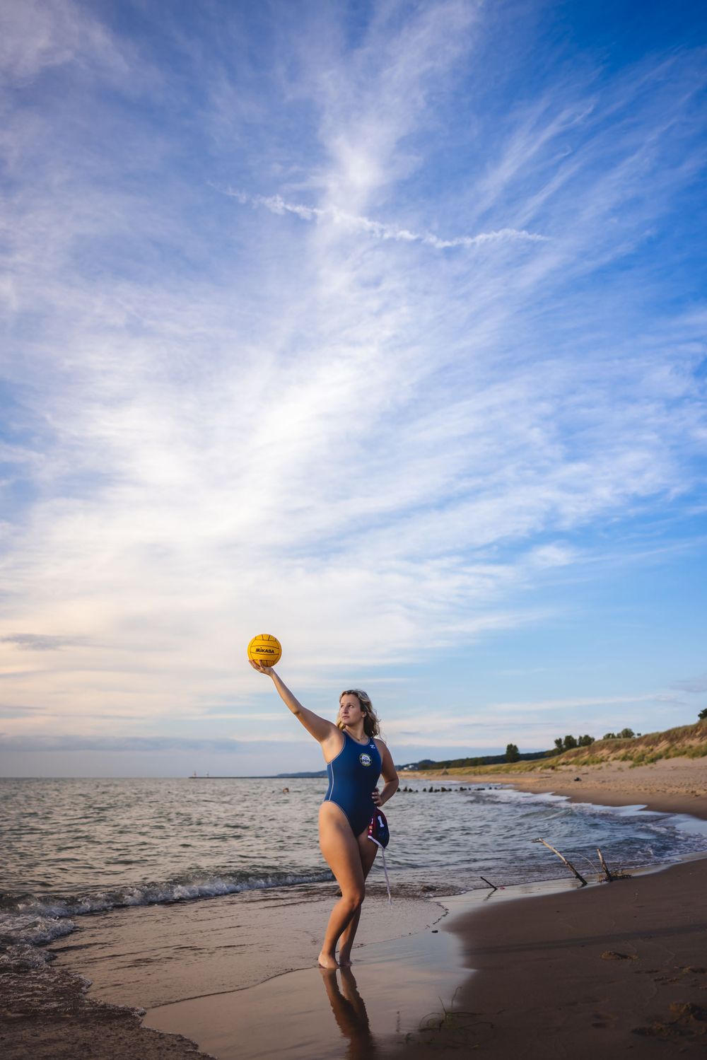 Water polo player jumps with ball against dramatic cloudy sky on beach at sunset.