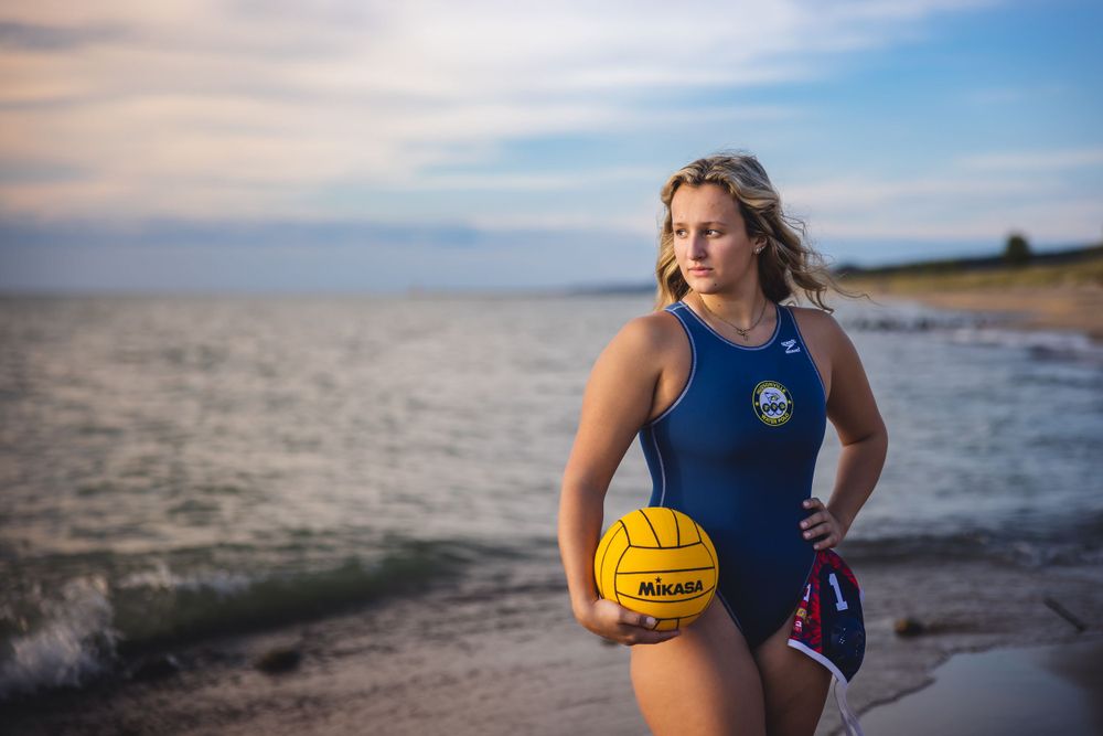 Athletic figure in navy swimsuit holds yellow water polo ball on beach at sunset.