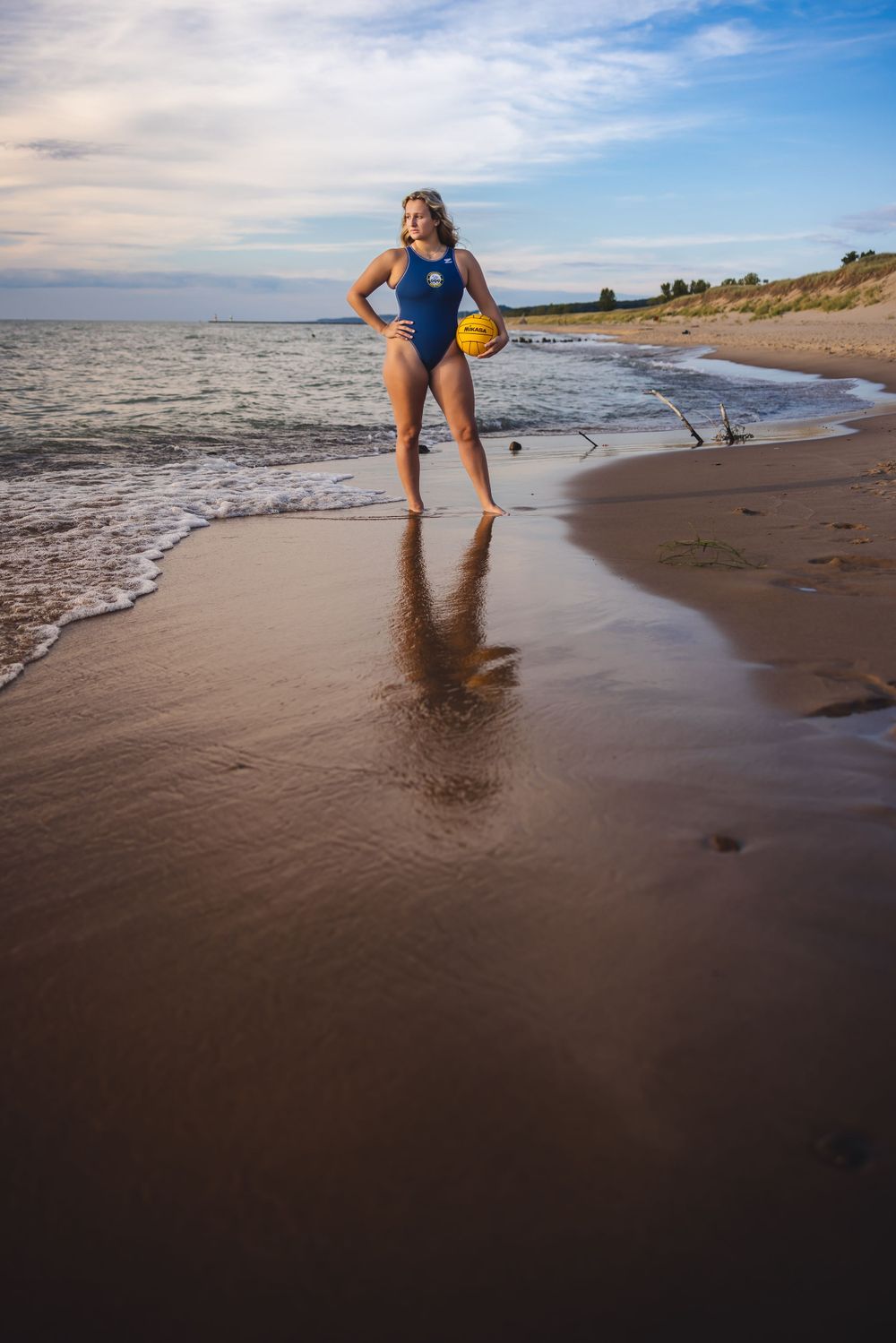 Water polo athlete poses with yellow ball on wet sand with waves rolling in.