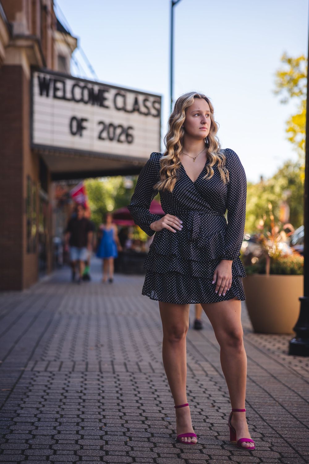 Senior portrait in front of welcome class of 2026 sign with stylish polka dot dress and pink heels.