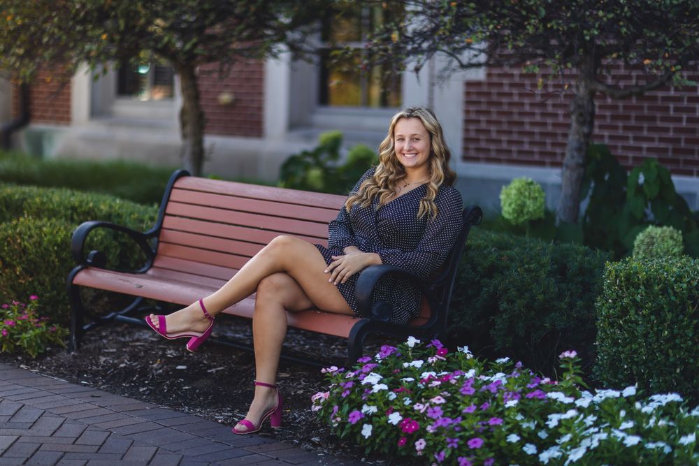 Senior portrait on park bench surrounded by flowers wearing navy dress and pink shoes.