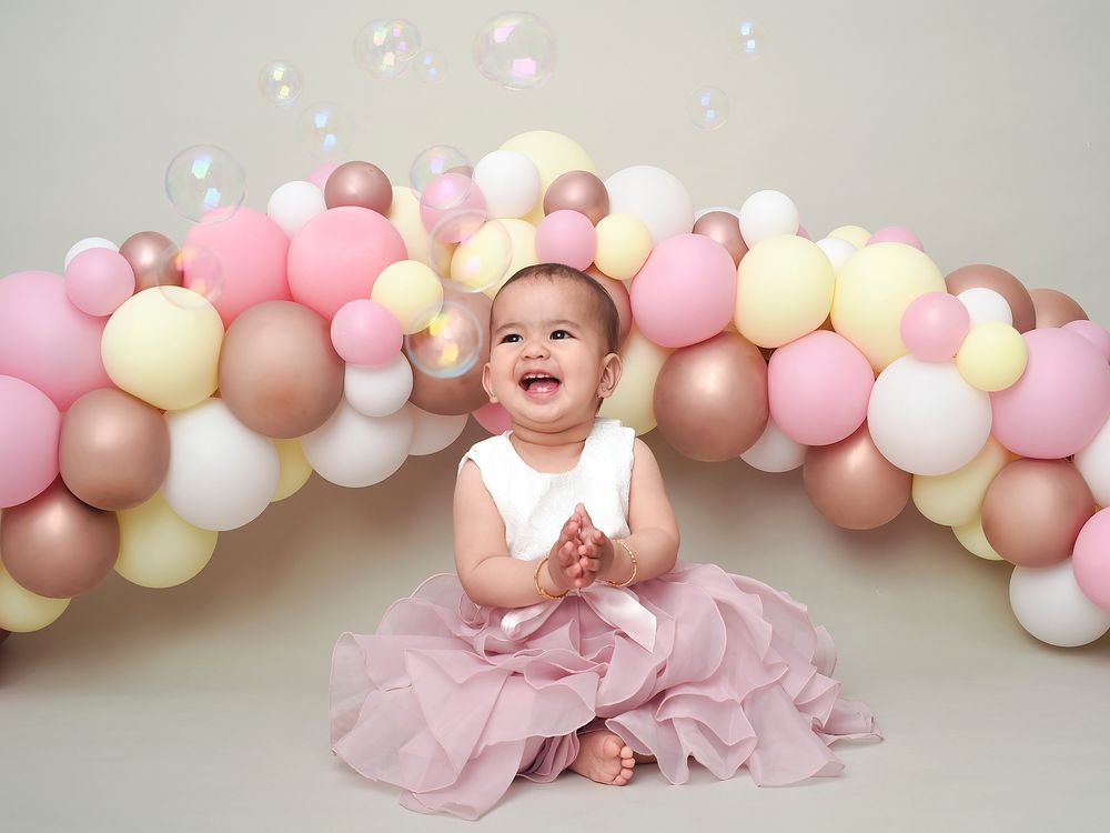 A baby girl in a pink dress sits in front of colorful balloons during her one-year photography session at Abigail's studio.