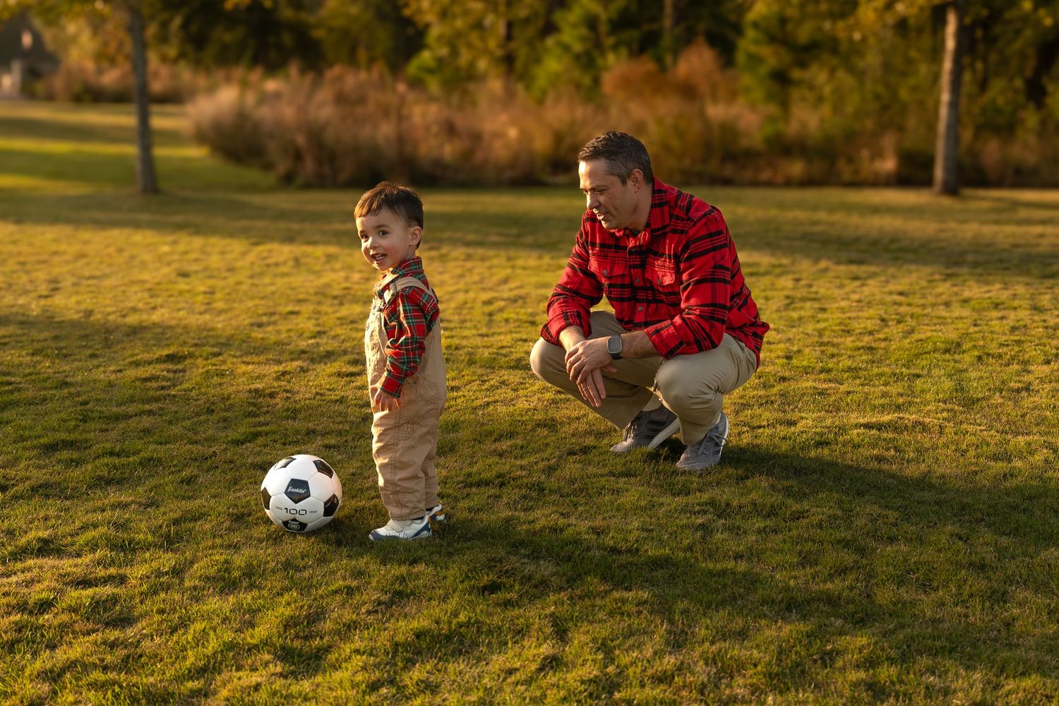 Adult and child playing soccer together on a grassy field during golden hour sunset.