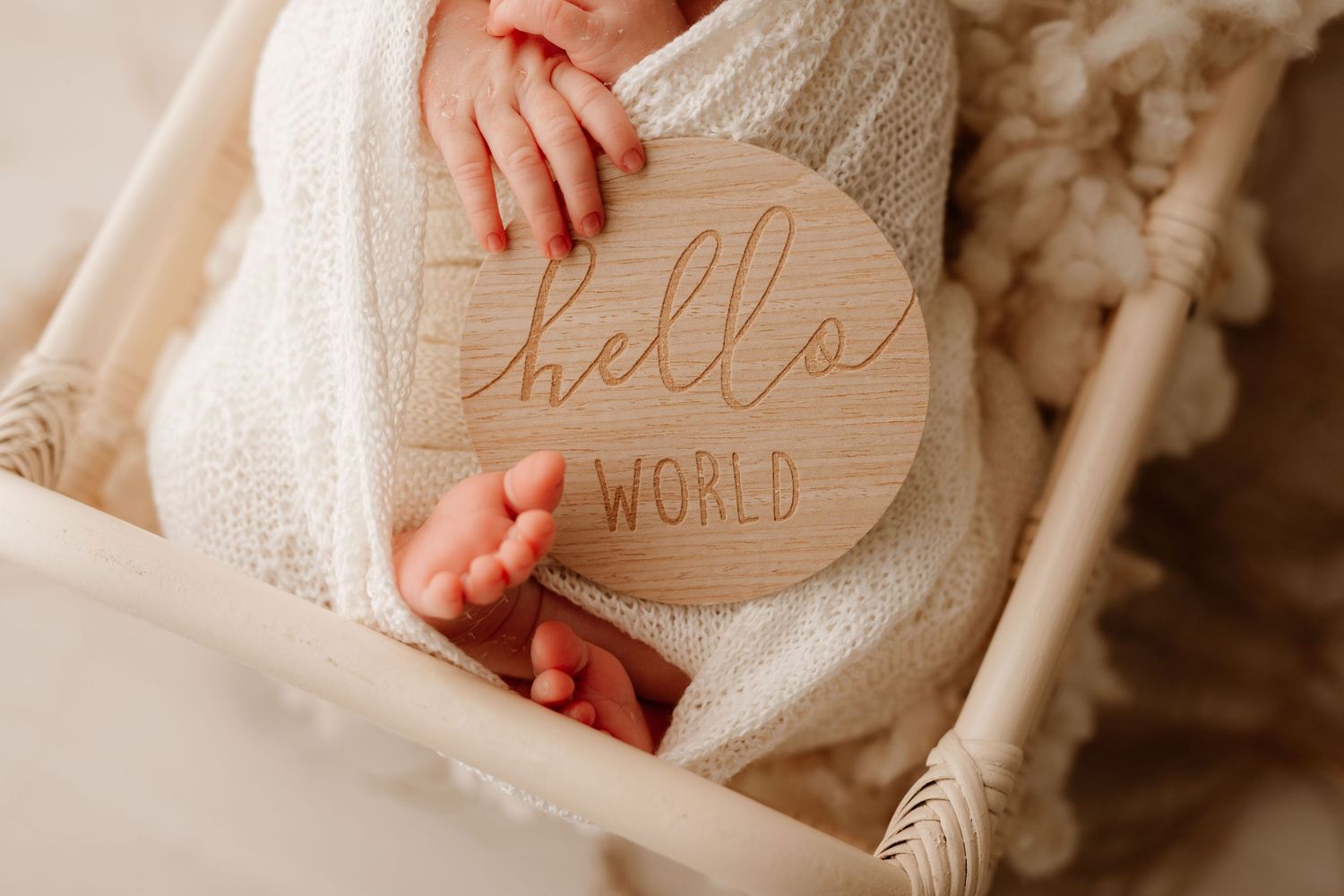 Newborn baby wrapped in white swaddle holding a wooden Hello World sign in a boho-style crib.
