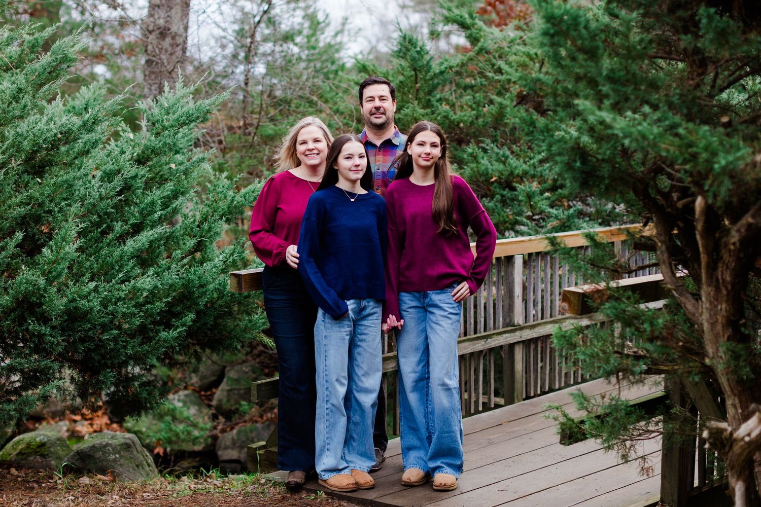 A group of four people in coordinating burgundy and blue outfits pose together on a wooden bridge in a forest setting.