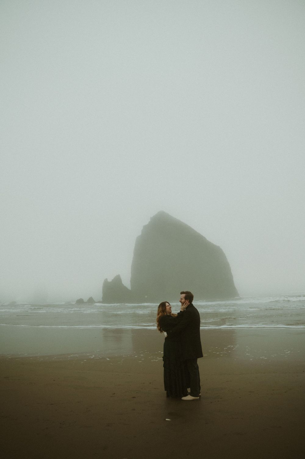 Silhouettes on a misty beach with large rock formations visible in the foggy background.