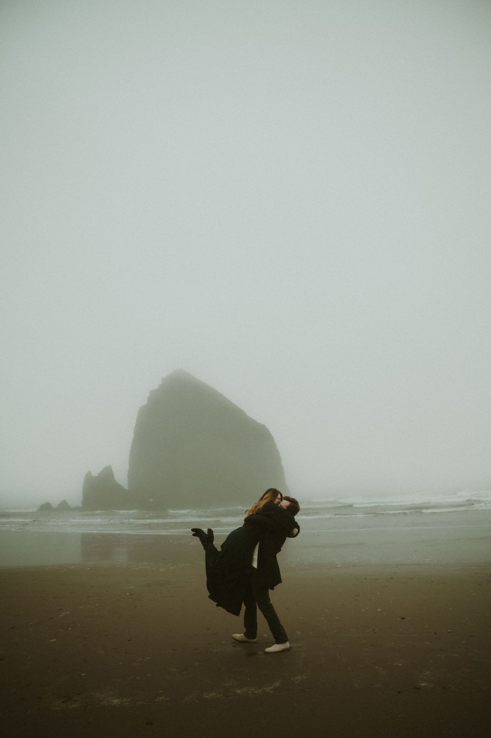 Figures in dark clothing moving across a foggy beach with sea stacks in the distance.