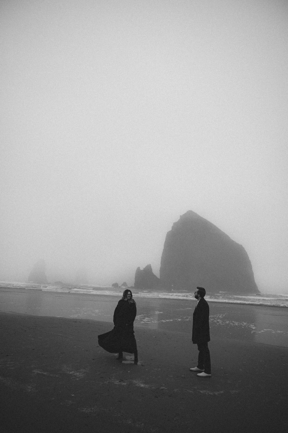 Black and white photo of figures standing on a misty beach with Haystack Rock in the distance.
