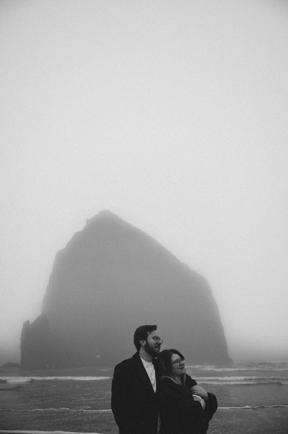 Silhouettes of large rock formations emerge from dense fog along a misty coastline.