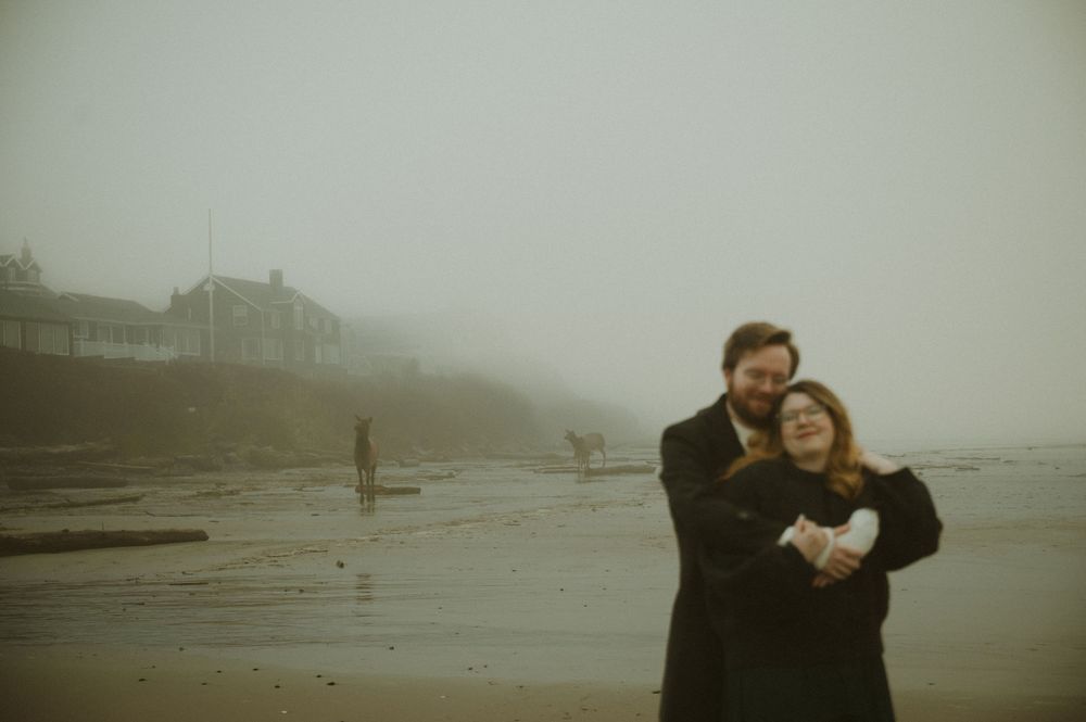 Romantic couple embraces on a misty beach with dark, moody atmosphere and foggy weather in the background.