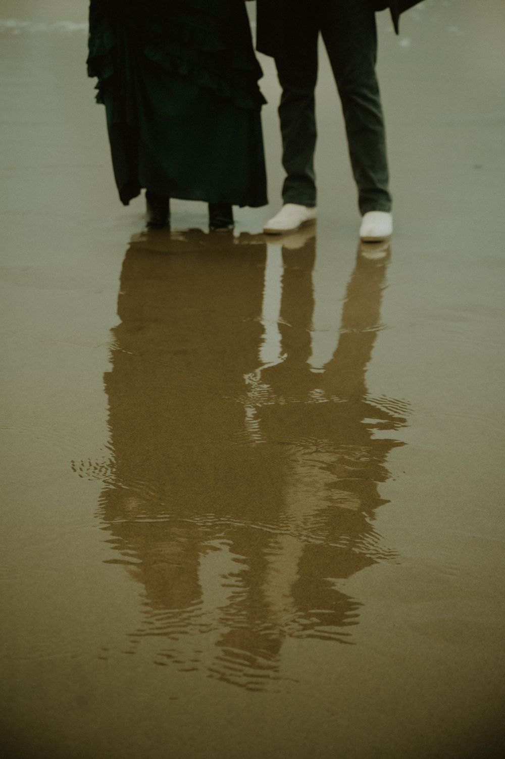 Reflections of people's legs and feet in wet sand on an overcast beach.