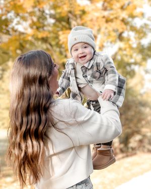 Mother in white sweater lifting and playing with young child in autumn setting.