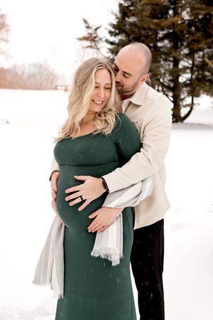 A couple embraces lovingly in a snowy winter setting wearing green and cream colored outfits.