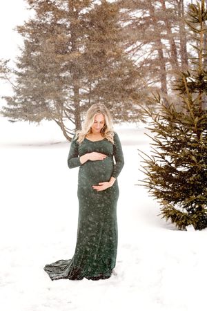 A woman in a dark green dress poses in a snowy winter landscape with evergreen trees in the background.
