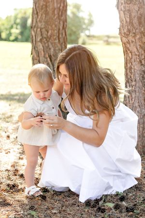A tender moment captured in sunlight as a parent and toddler explore something together under pine trees.