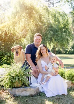 A family and their golden retriever pose together on a garden stone surrounded by lush ferns on a sunny day.