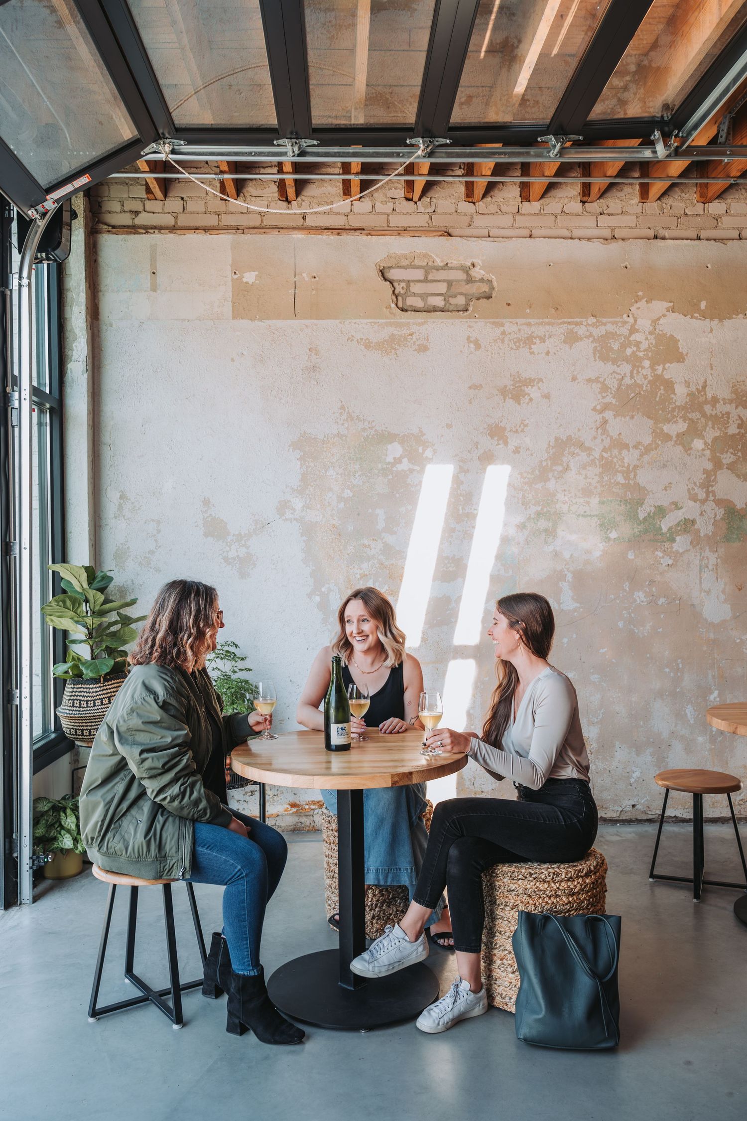 Two people sit and chat on woven ottomans at round wooden tables in an industrial-style cafe space with exposed brick walls.