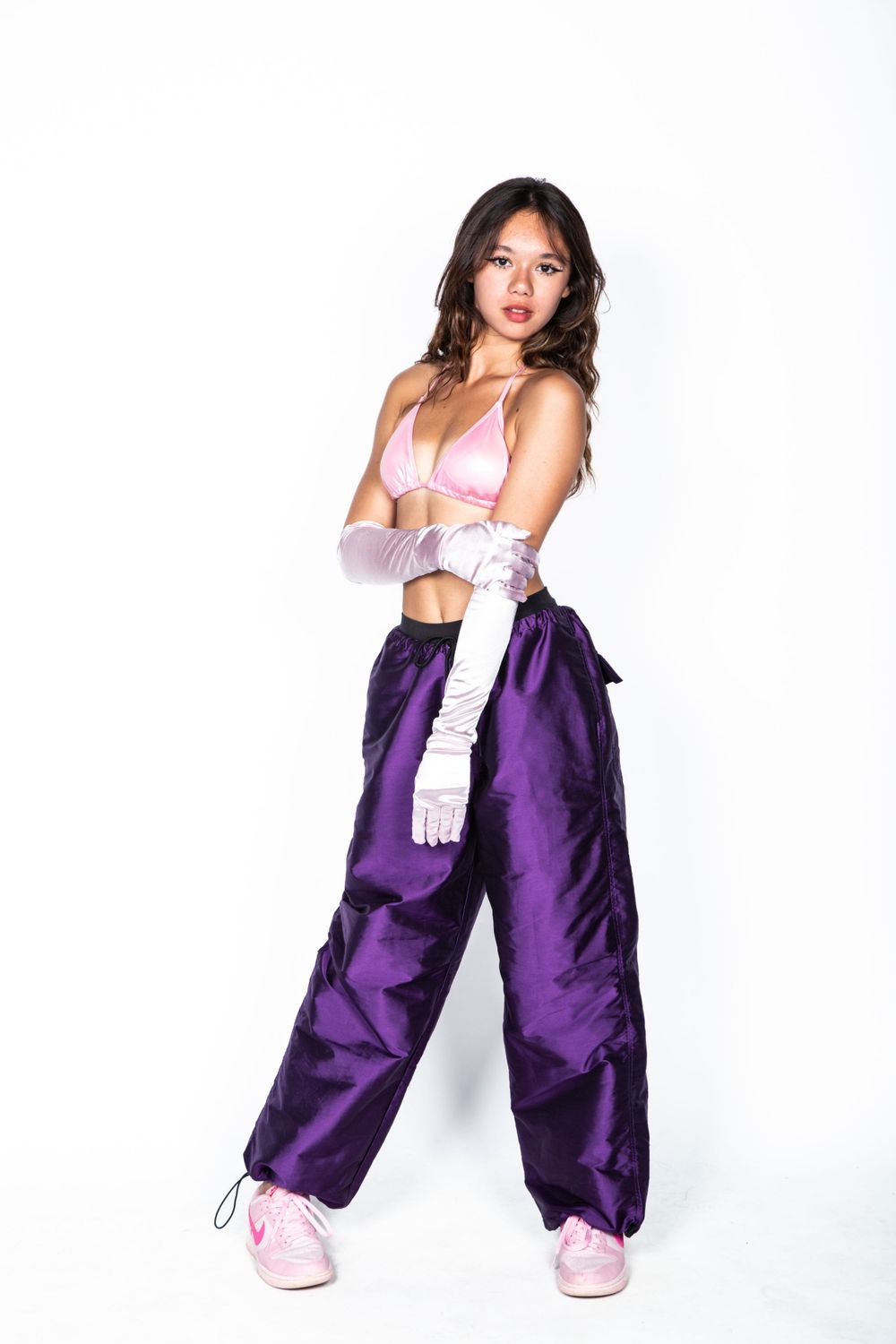 Athletic figure in pink sports bra, white gloves and purple track pants poses against white background.