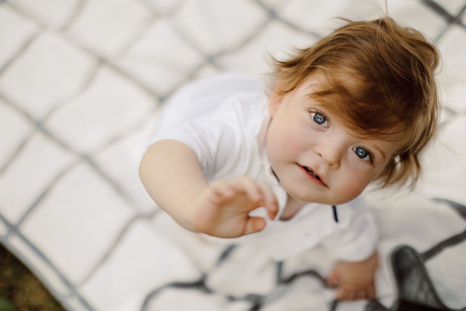 Playful toddler reaching toward camera on patterned blanket.