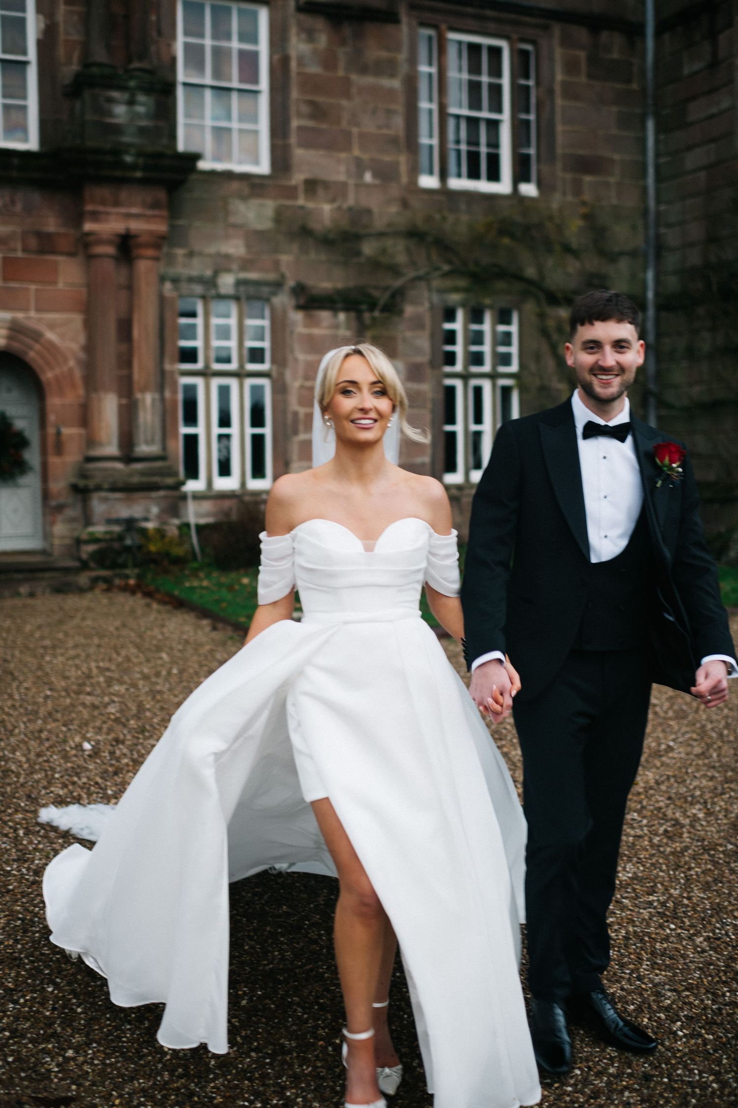 A couple in wedding attire playfully runs together on a gravel courtyard outside a stone building. Editorial wedding photographer.