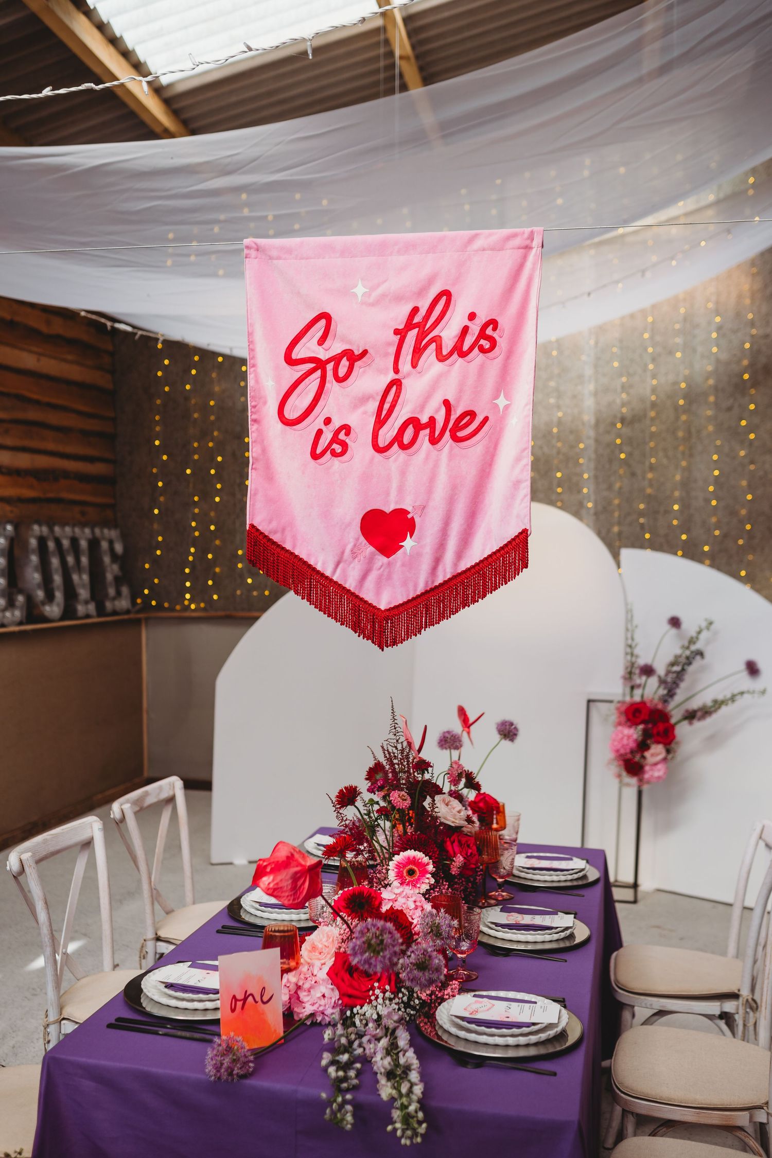 Modern wedding reception table setup with purple tablecloths, pink banners, and floral centerpieces under string lights.