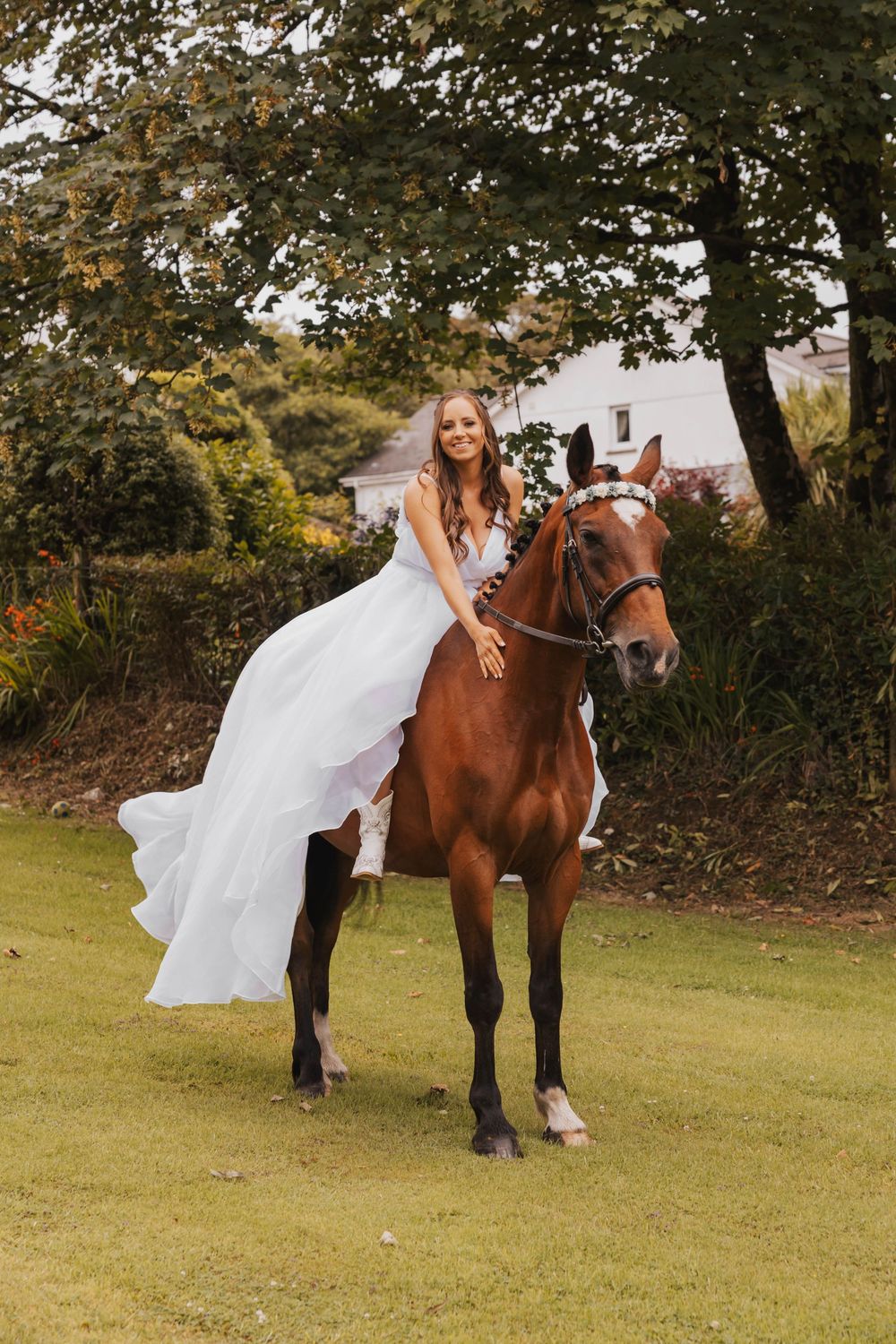 A beautiful chestnut horse stands on grass while carrying someone in a flowing white dress during an outdoor photo session.