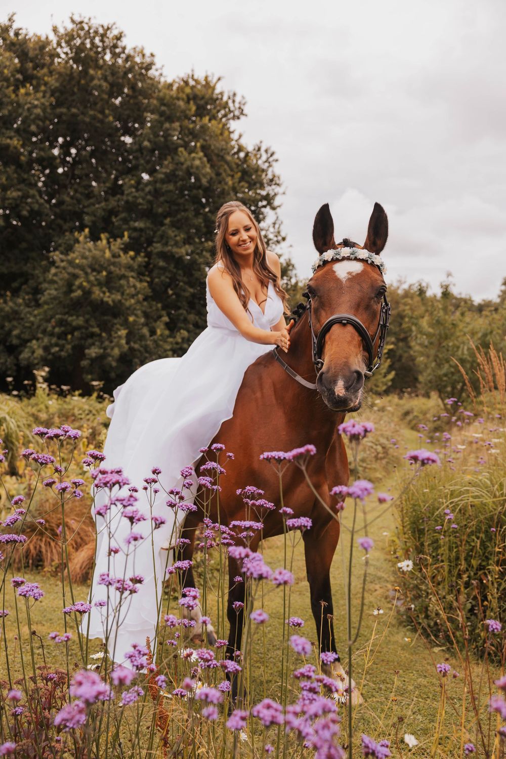 A white dress contrasts beautifully against purple wildflowers and a chestnut horse in a dreamy outdoor meadow setting.