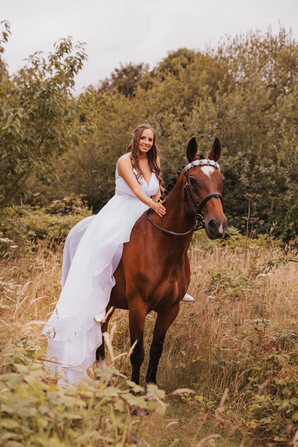 A person in a flowing white dress poses with a brown horse in a sunlit meadow filled with tall grass and trees.