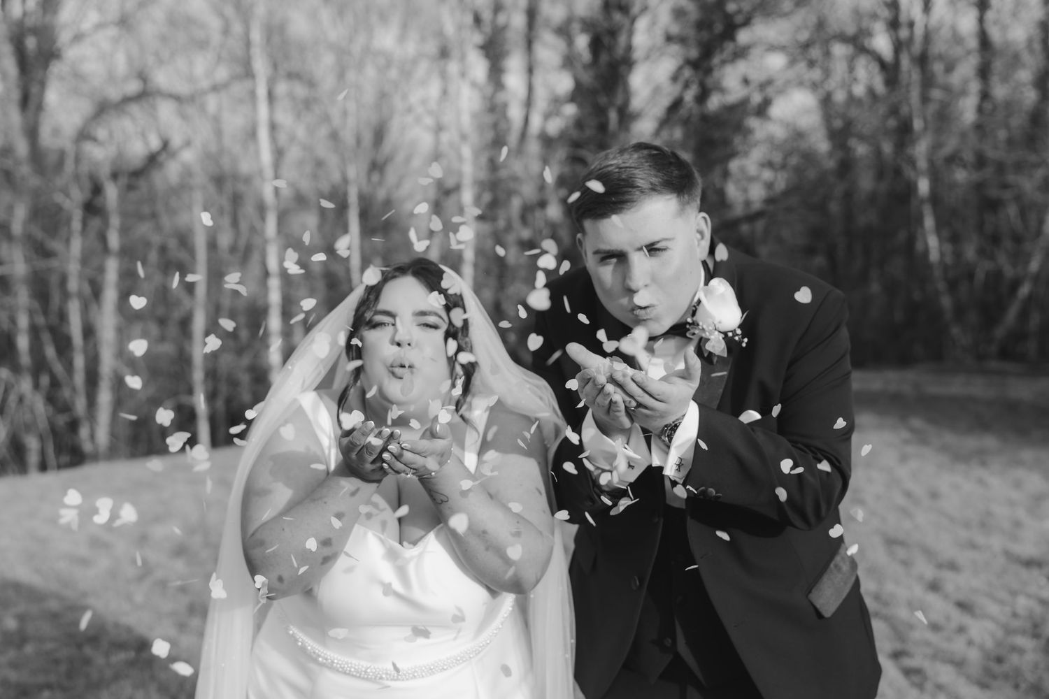A black and white wedding photo of a bride and groom playfully blowing confetti at the camera outdoors.