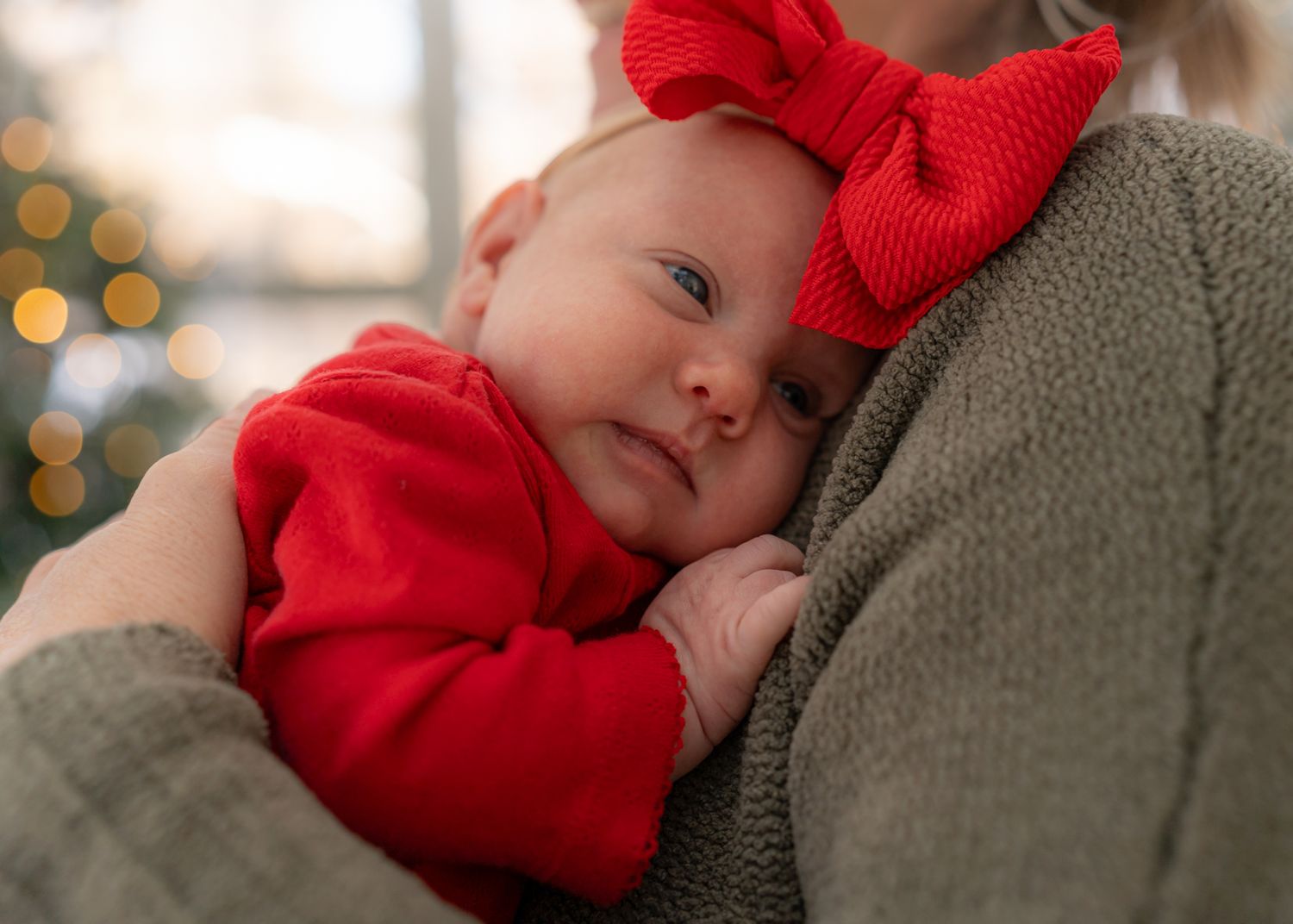Baby in red outfit rests against cozy gray sweater in warm indoor lighting.