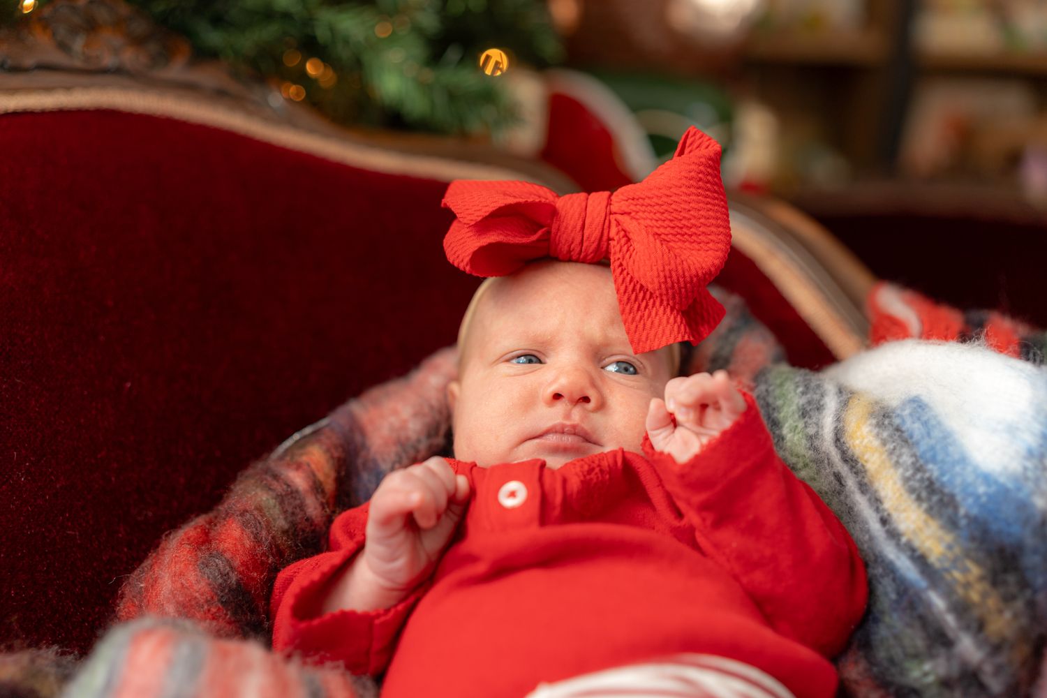 Baby wearing bright red outfit rests on patterned blanket with holiday lights in background.