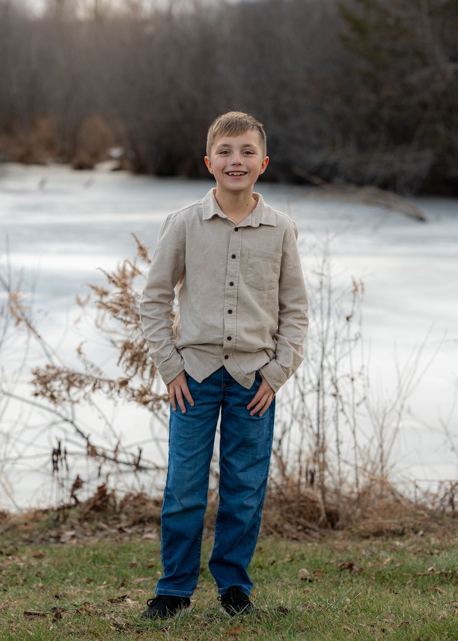 Child in beige button-up shirt and blue jeans standing in snowy winter landscape.