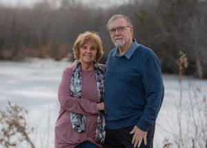 A couple poses together by a frozen lake on an overcast winter day wearing blue and mauve sweaters.