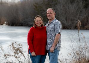 A couple stands together outdoors by a frozen lake, with her in a red sweater and him in a patterned shirt.