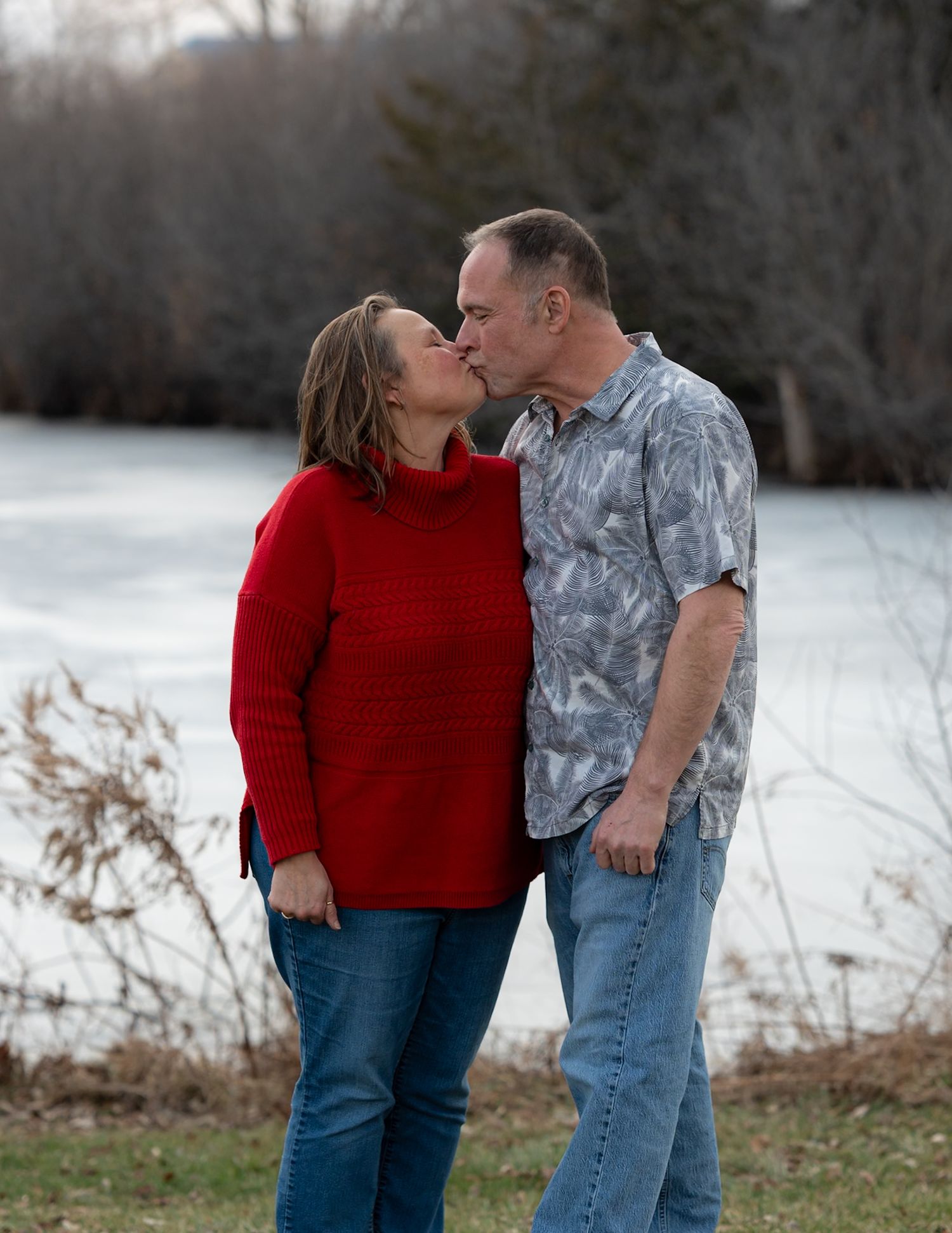 A romantic moment captured between a couple by a frozen lake, with her in red and him in casual attire.