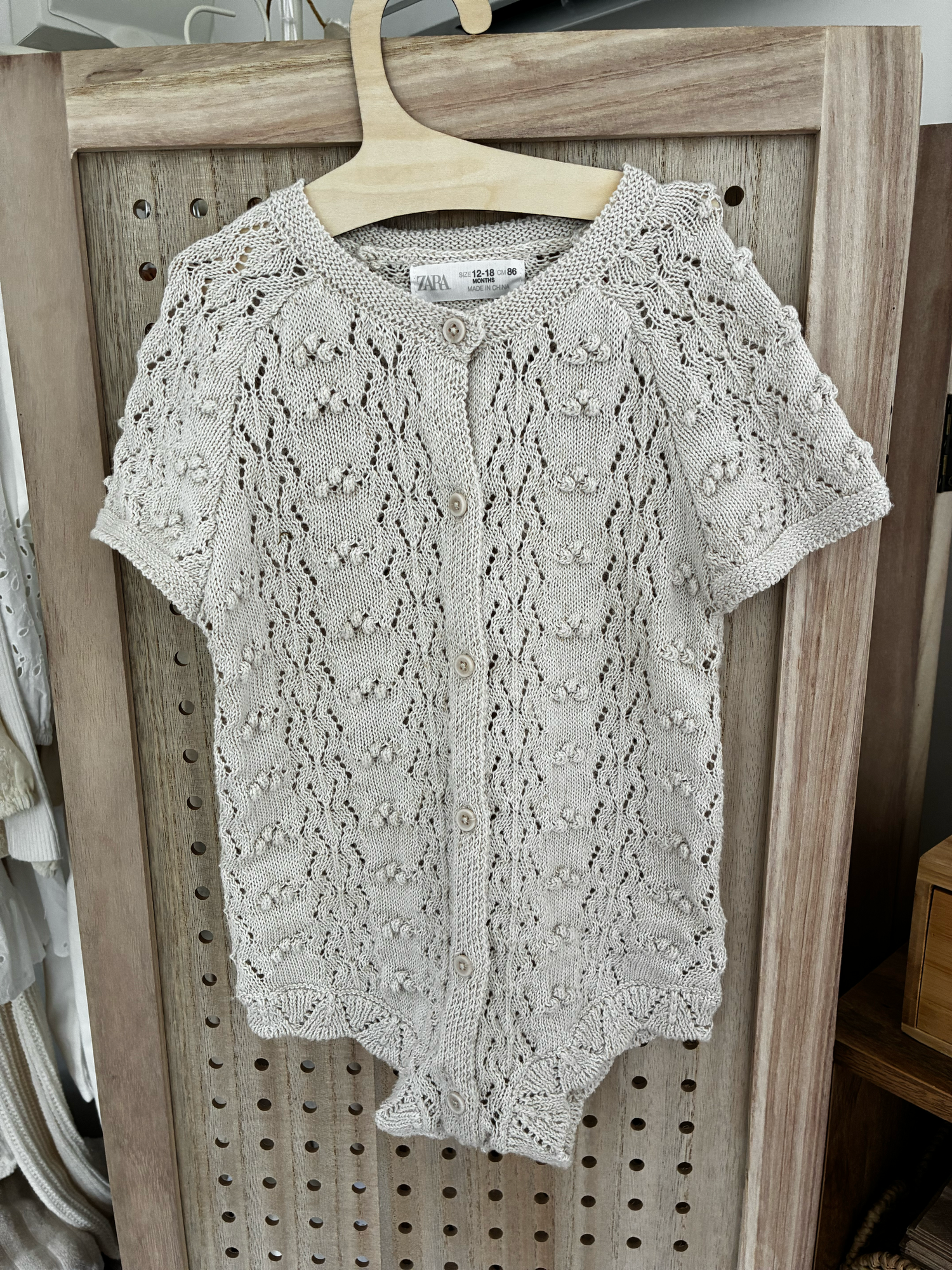 Vintage-style beige knitted baby romper with button details and side pockets hanging on display.