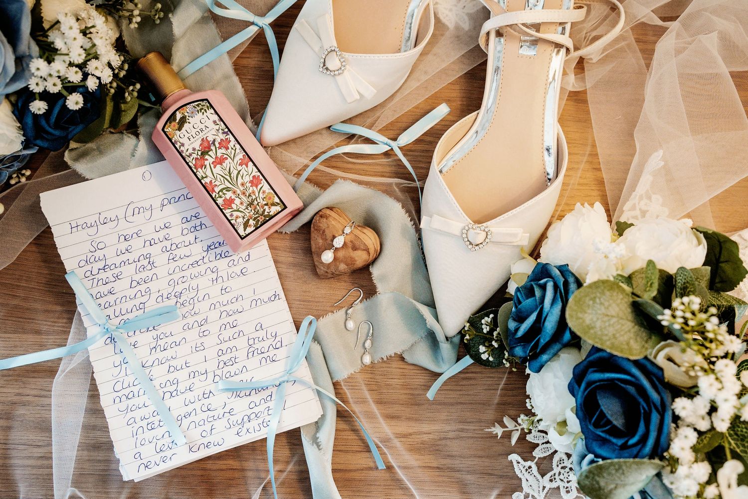 Wedding details including handwritten notes, blue flowers, and bridal accessories arranged on wooden surface.