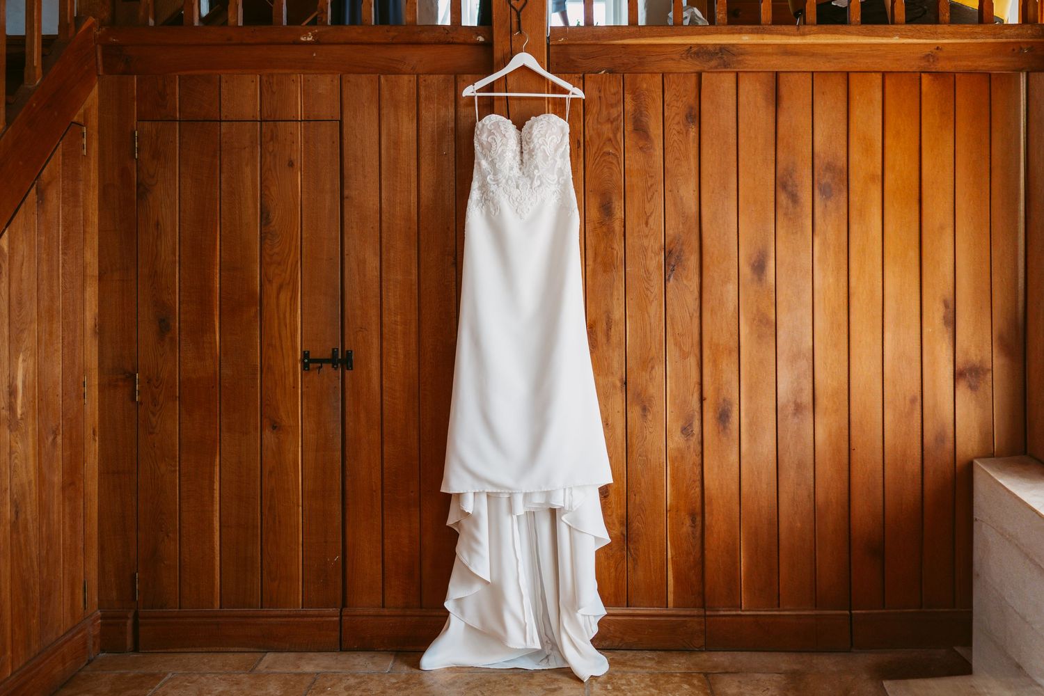A white wedding dress hangs against wooden paneled walls in warm lighting.