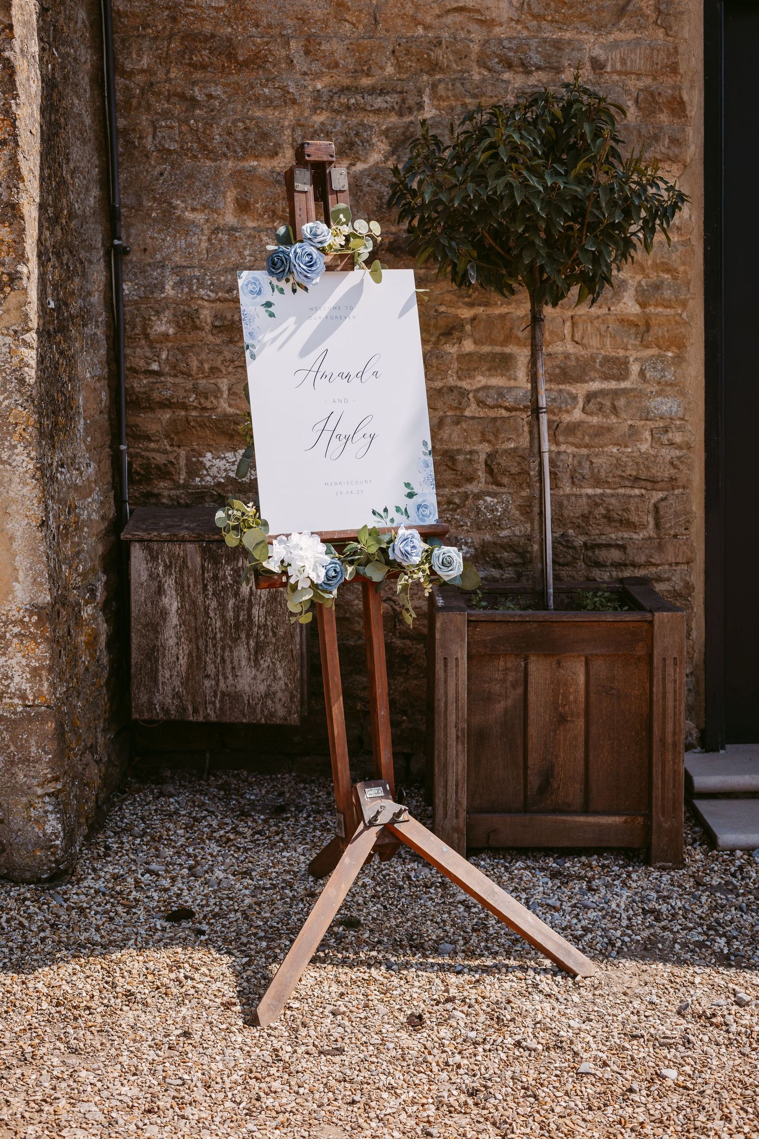 Wooden signs with floral decorations stand on gravel against a rustic stone wall backdrop.