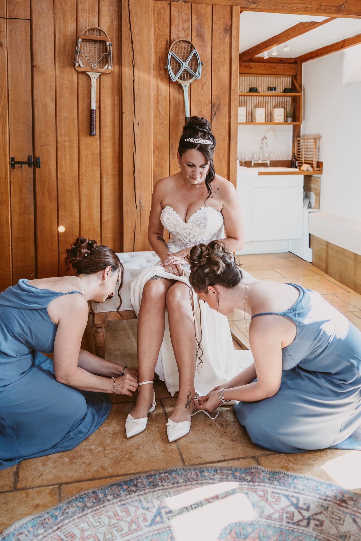Bridesmaids in blue dresses helping with wedding shoe fitting in rustic wooden room.