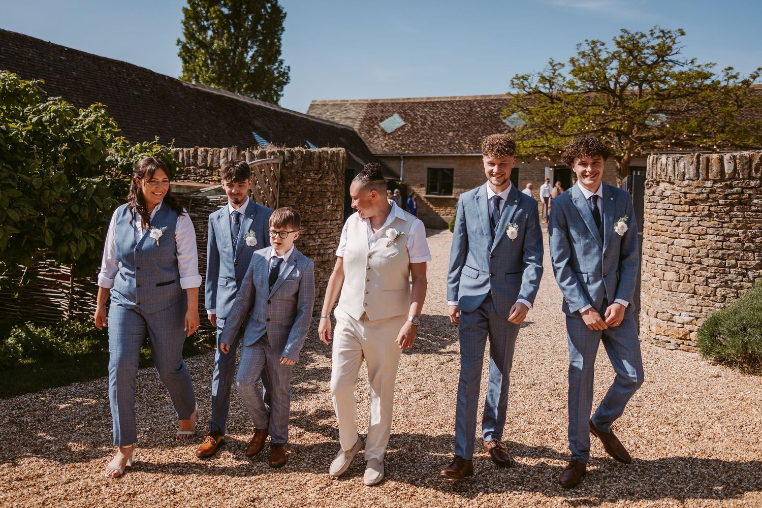 Wedding party in blue and cream suits walking together on a gravel path outside a rustic stone building.