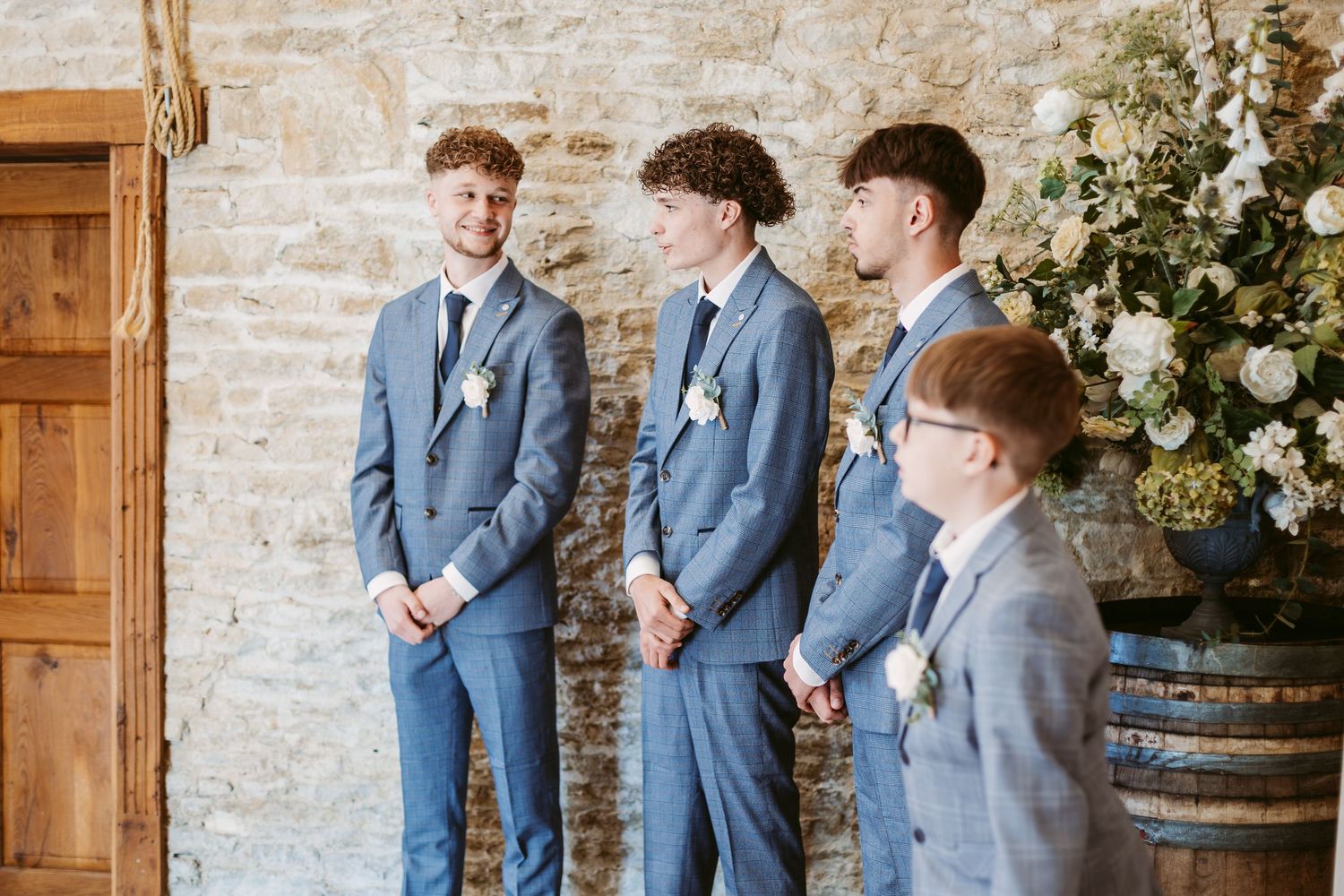 Groomsmen in blue suits stand together in front of rustic barn wall.