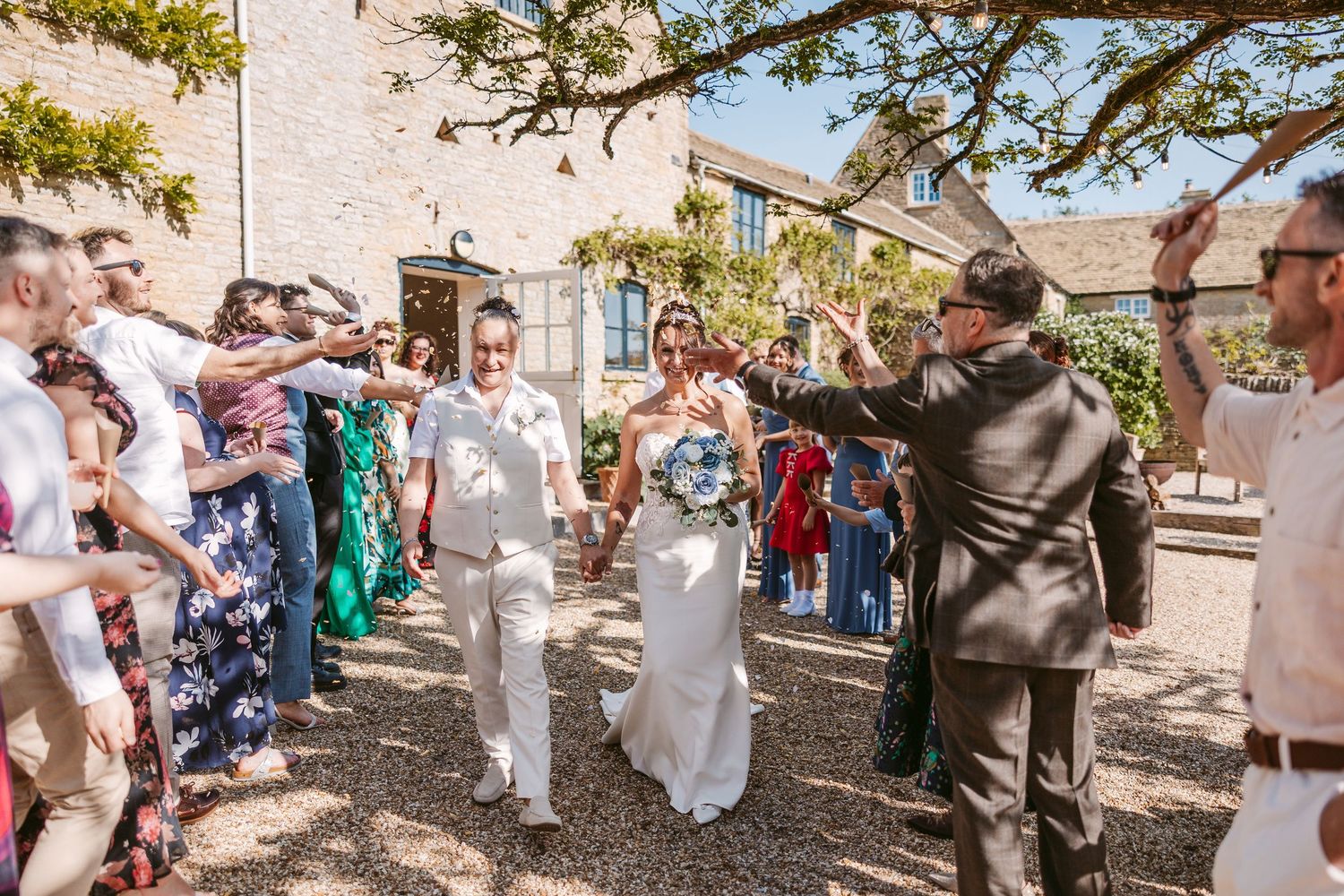 Wedding guests celebrate outdoors under trees with confetti and joyful expressions.